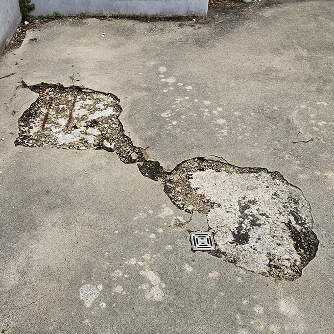 Asphaltboden mit zwei unregelmäßigen, verbundenen Ausbesserungsstellen und einem kleinen Metallablauf in der Mitte